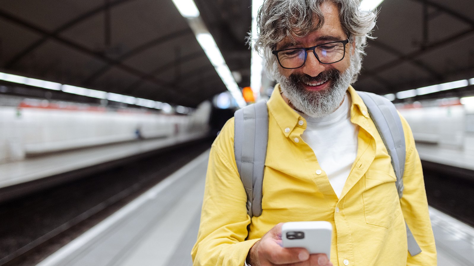 Älterer Mann am Smartphone im gelben Hemd und Rucksack am Bahnsteig