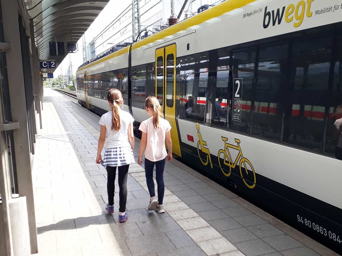 Zwei junge Mädchen stehen mit dem Rücken zur Kamera am Bahngleis neben einem Zug.