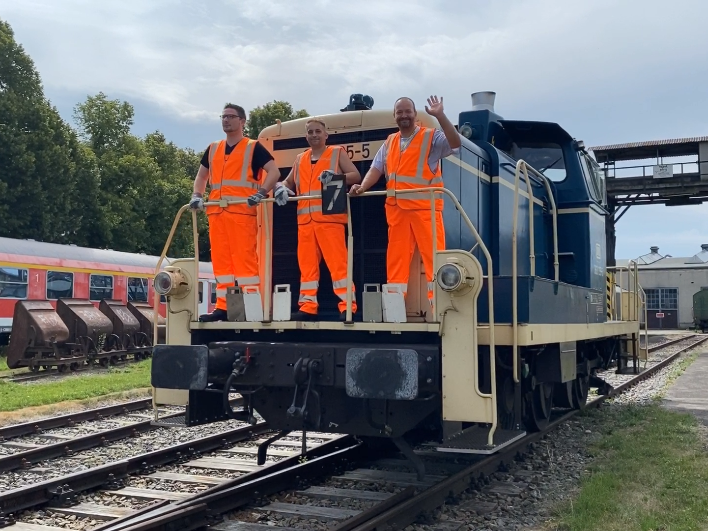 Drei Triebfahrzeugführer stehen auf einer Bahn und winken in die Kamera.