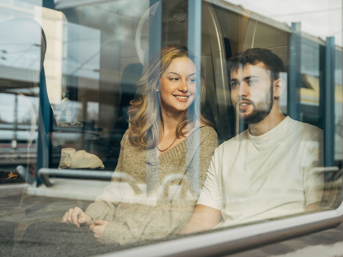 Junge Mann und Frau sitzen nebeneinander im Zug. Sie sind durchs Fenster zu sehen.