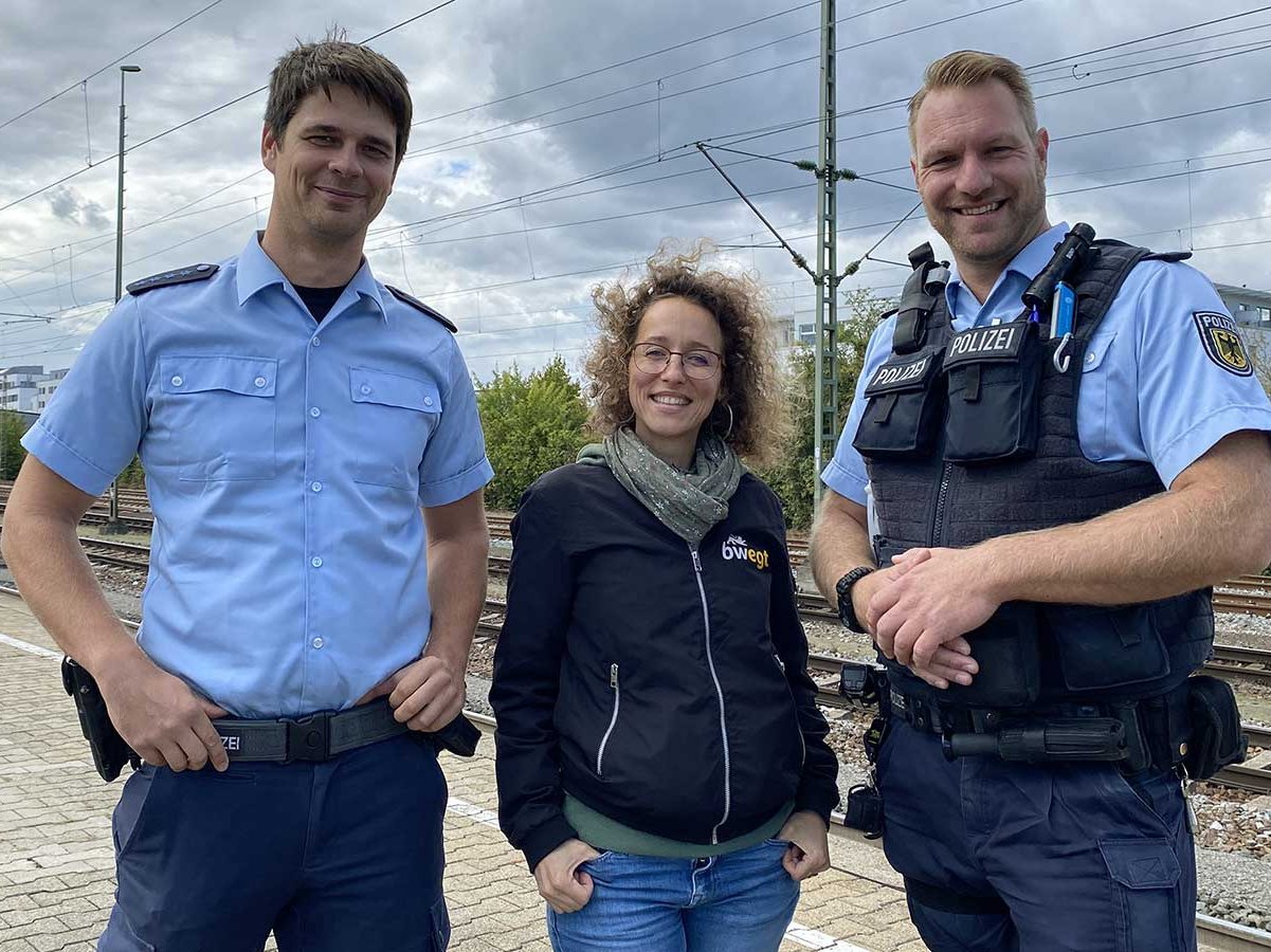 Frau steht zwischen zwei Polizisten am Bahngleis.