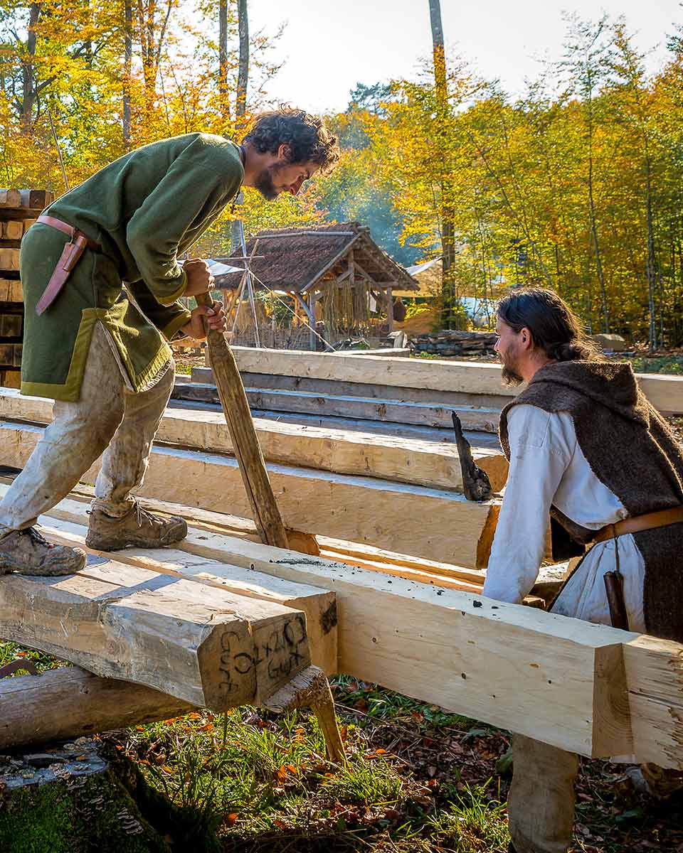 Zwei Handwerker in mittelalterlicher Kleidung schieben zuvor bereits zersägte Holzstämme zusammen.