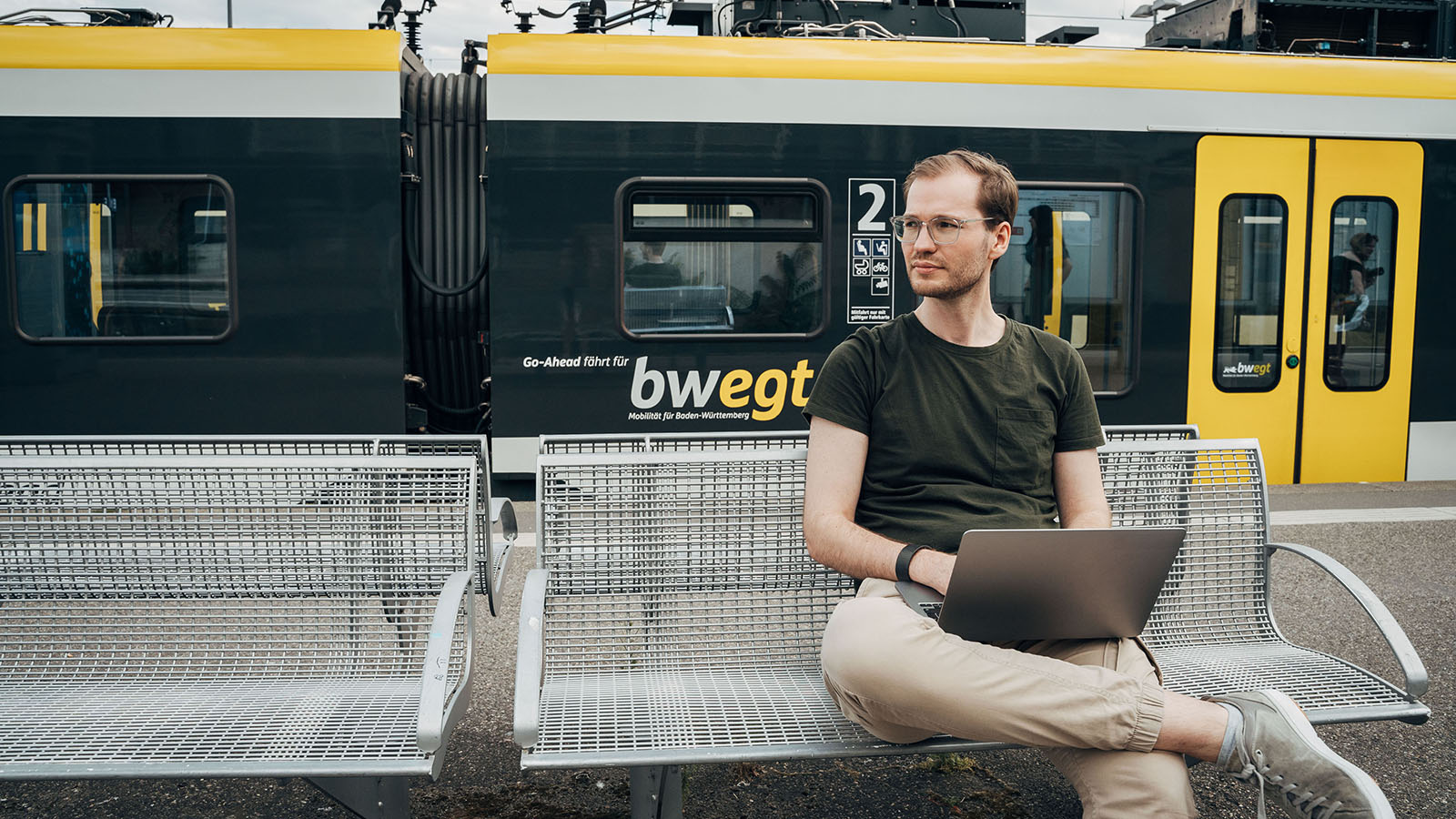 Ein Mann sitzt am Bahnhof mit seinem Laptop und im Hintergund ist ein bwegt Zug zu sehen.