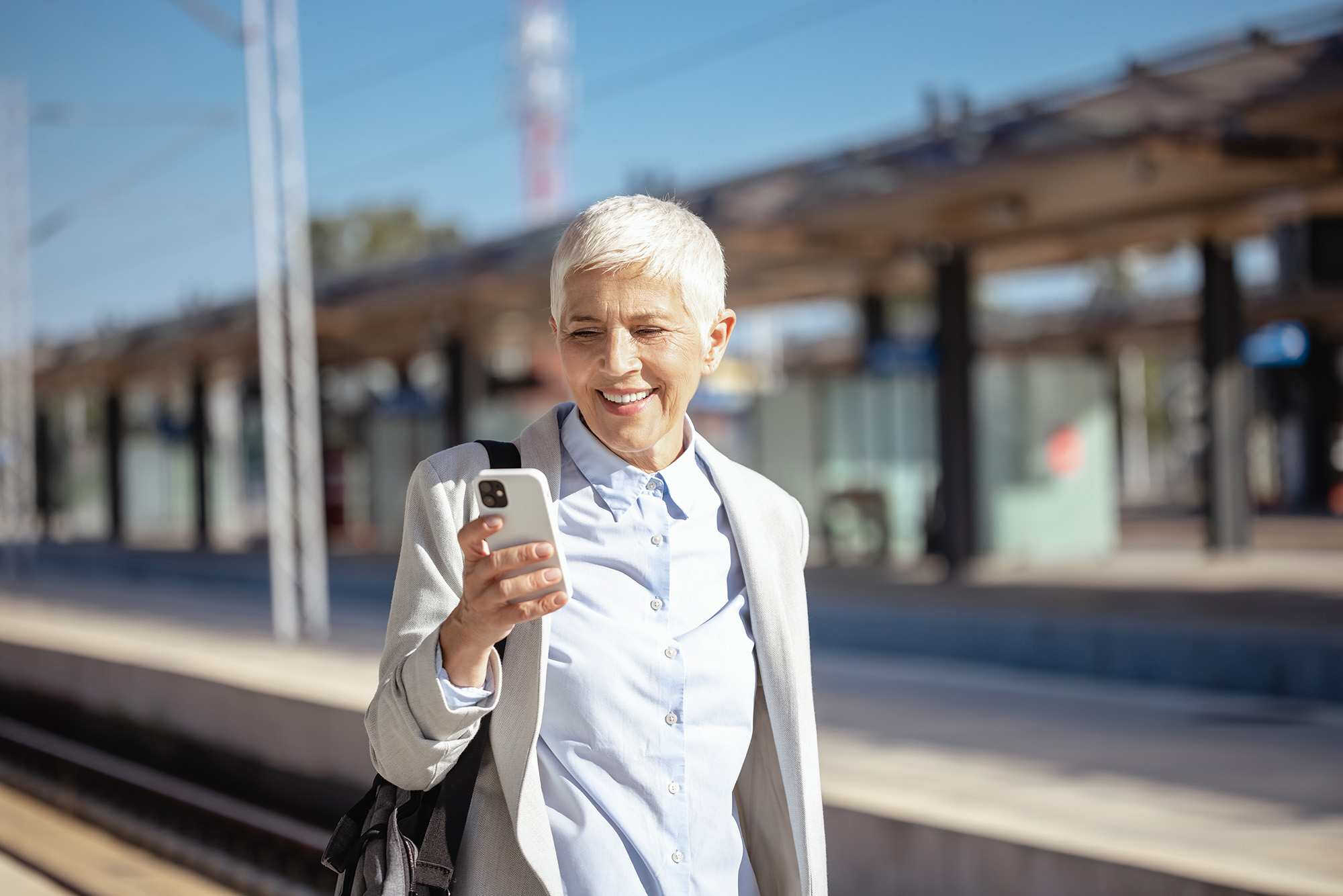 Eine Frau im Businessoutfit und weißen, kurzen Haaren schaut am Bahnsteig auf ihr Smartphone