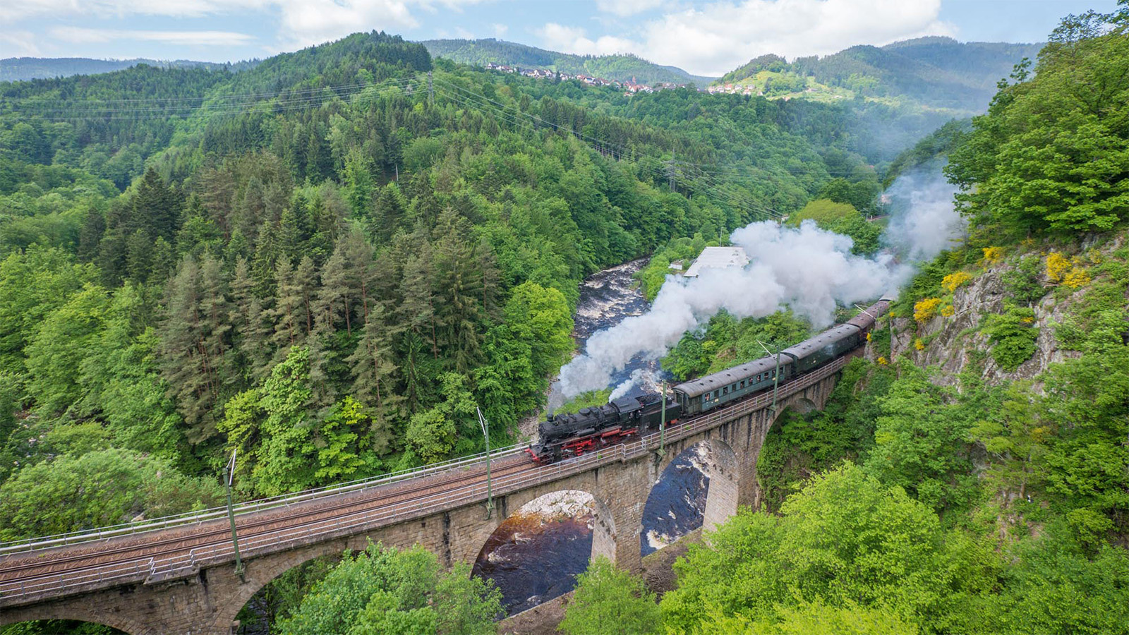 Eine rußende Dampflokomotive auf einer hohen Eisenbahnbrücke im bewaldeten Gebirge