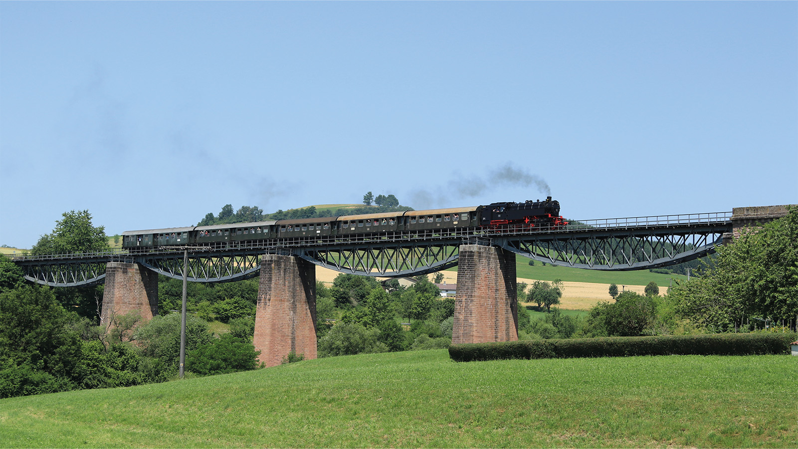 Eine rauchende Dampflokomotive, die durch grüne Landschaft über eine Eisenbahnbrücke fährt