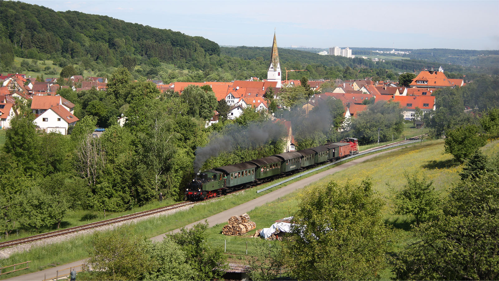 Panorma-Aufnahme einer dampfenden Lokomotive, die durch eine grüne, bergige Landschaft an einem kleinen, ländlichen Dorf vorbei fährt