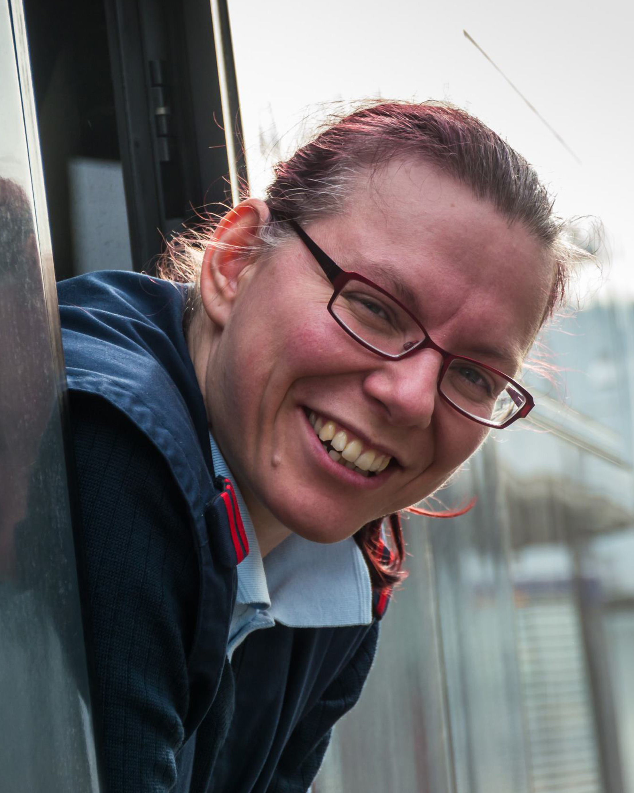 Lokführerin mit Brille lehnt sich aus Zugfenster und lacht in die Kamera.