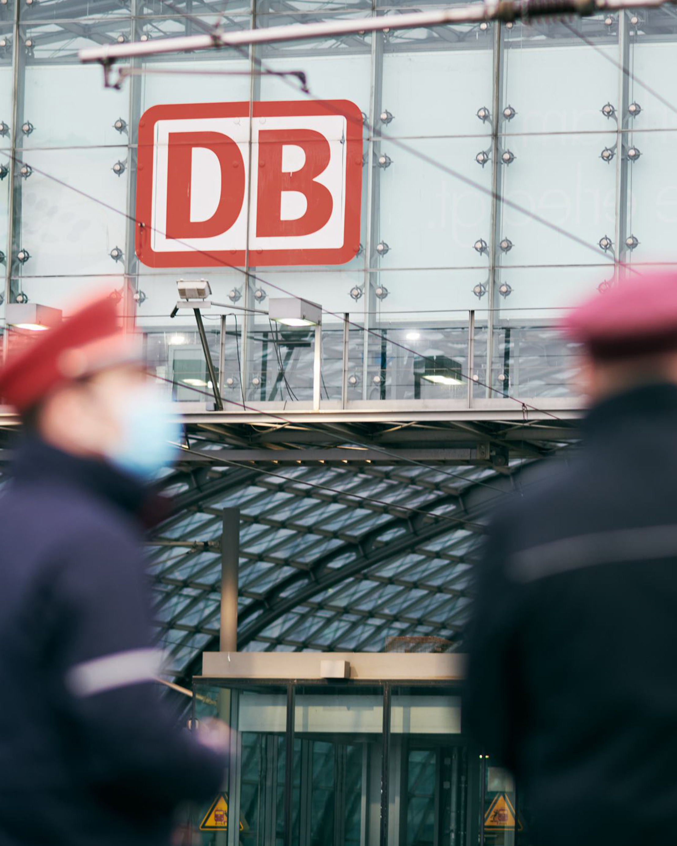 Bahnhof mit zwei unscharf zu sehenden Mitarbeitern im Vordergrund und dem DB-Logo im Hintergrund.