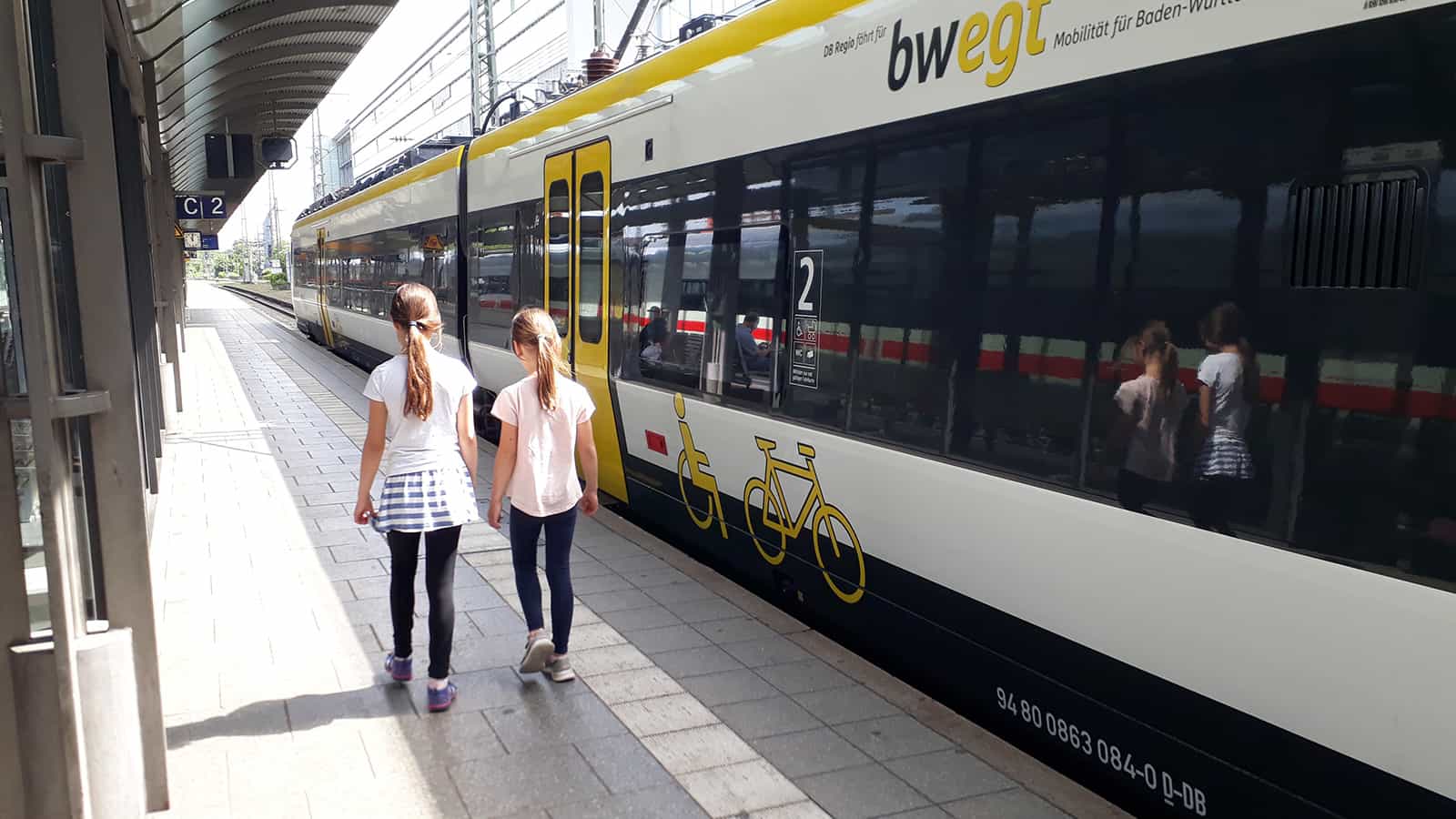 Zwei junge Mädchen stehen - den Rücken der Kamera zugewendet - am Bahngleis vor einem haltenden Zug.