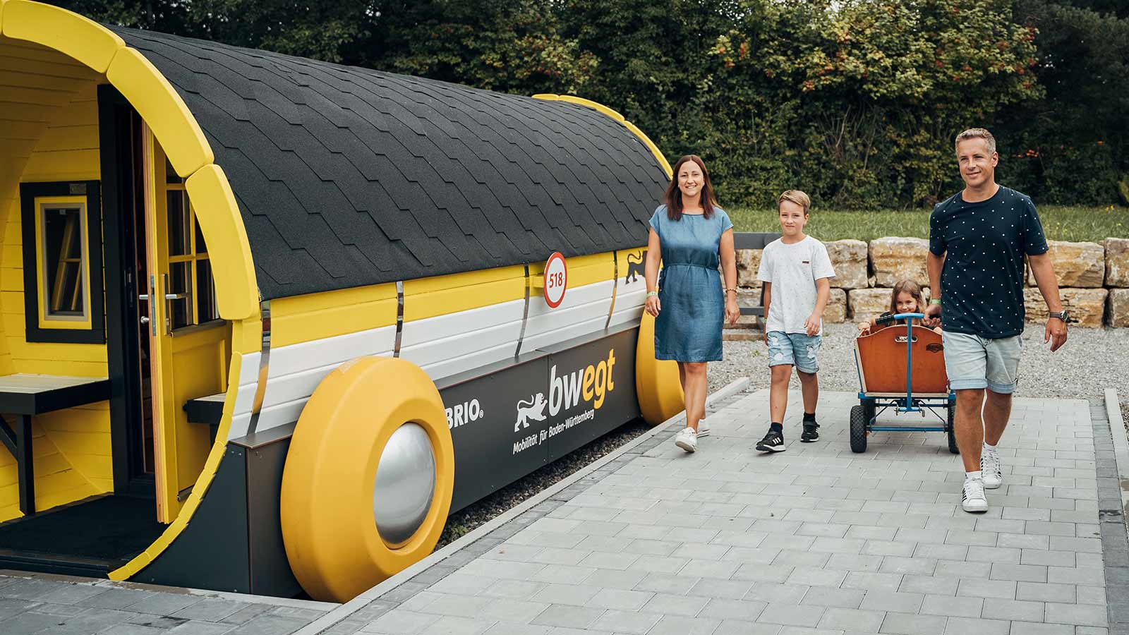 Familie mit zwei Kindern geht mit einem Bollerwagen an einem Brio-Eisenbahnwagon in bwegt-Farben vorbei.