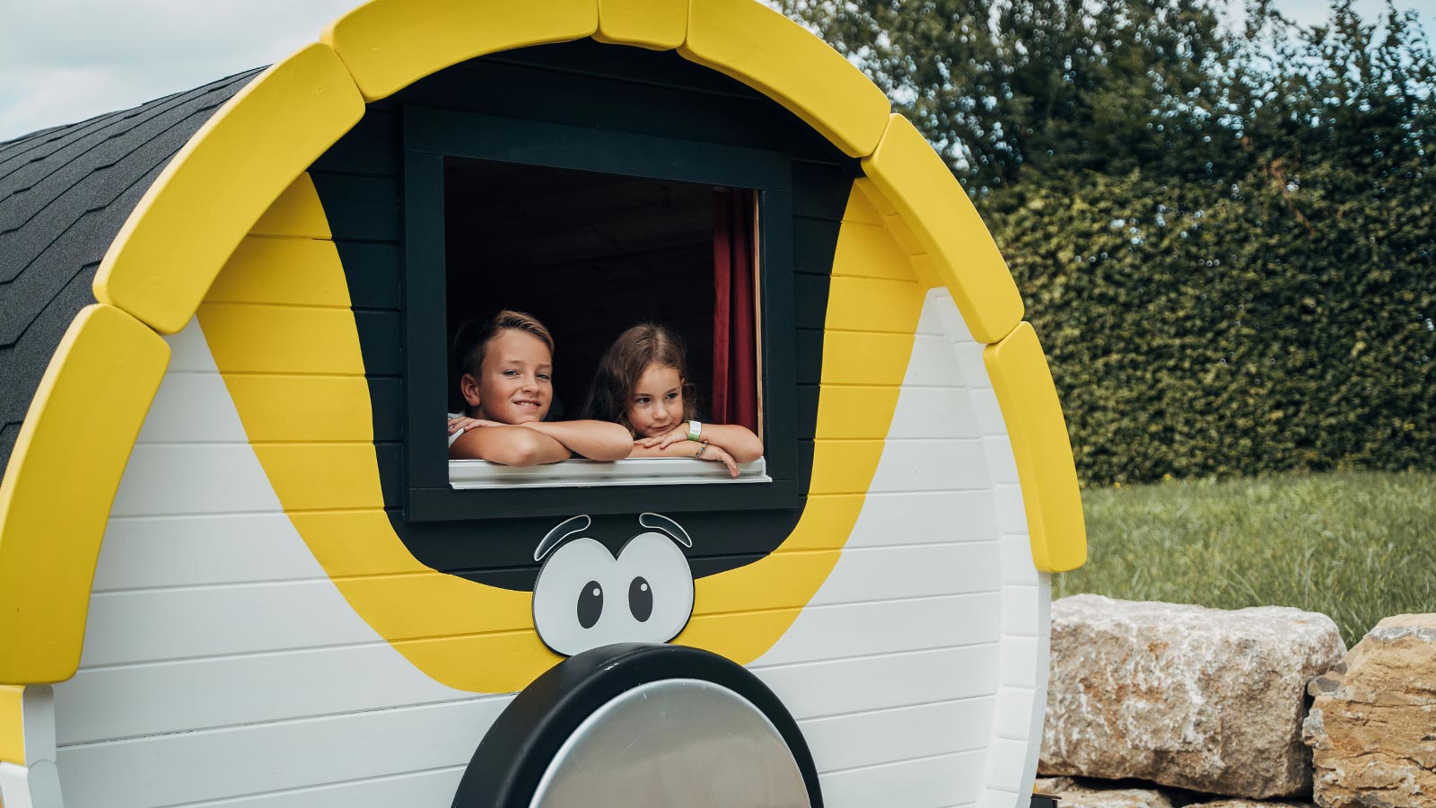 Zwei Kinder lehnen aus dem Fenster einer Holzhütte, die aussieht wie ein Zugwagon von Brio.