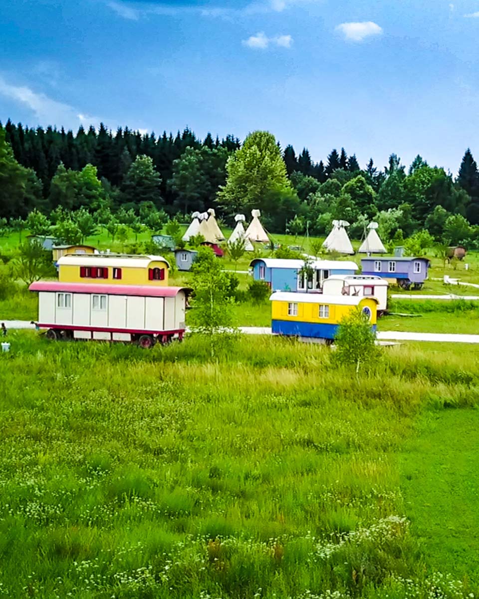 Zirkuswagen und Tipis stehen mitten auf einer Wiese umgeben von Wald.