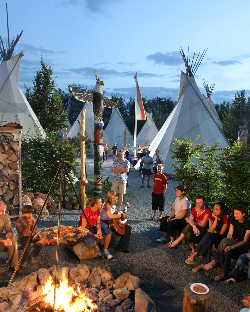 In einem Indianerdorf sitzen Menschen um ein Lagerfeuer herum. Im Hintergrund sind mehrere Tipis zu sehen.