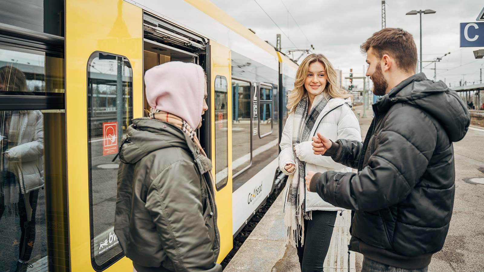 Drei junge Menschen sind mitten im Gespräch, während sie in eine Bahn einsteigen.