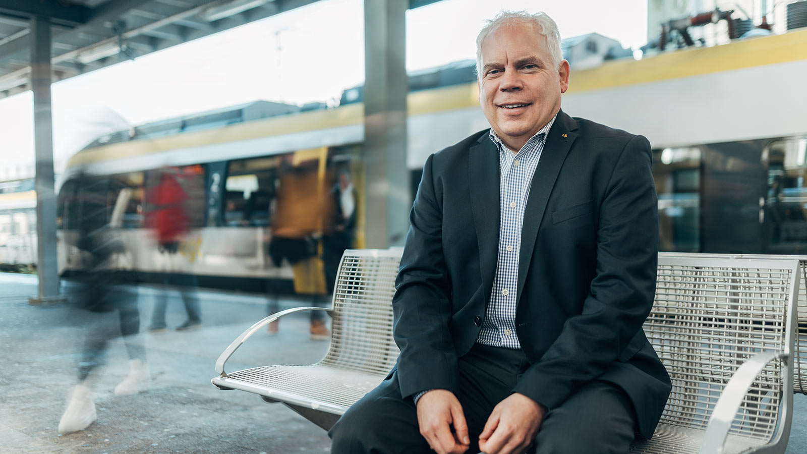 Mann sitzt am Bahnsteig auf einer Bank. Im Hintergrund steigen Fahrgäste aus einer Bahn.