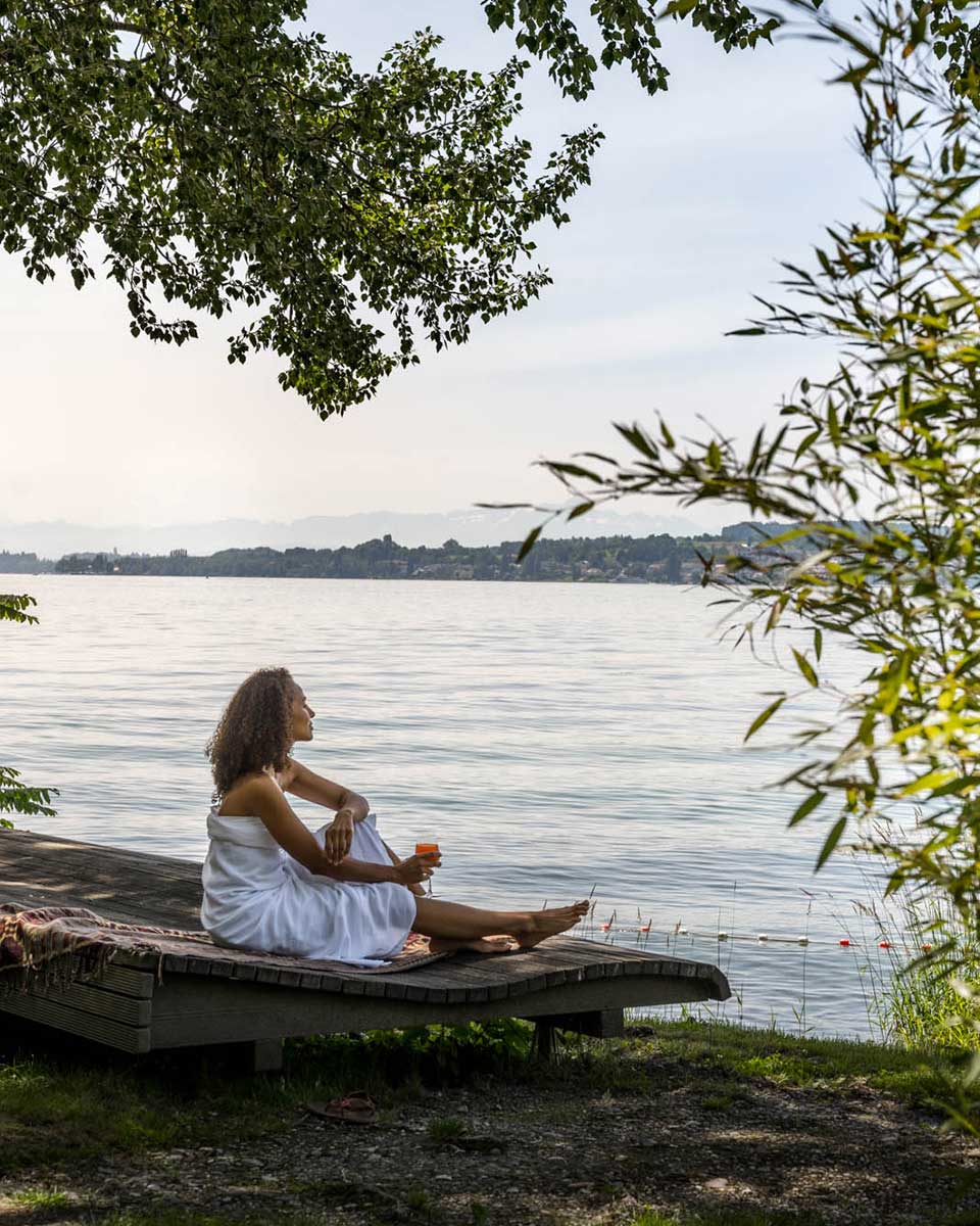 Junge Frau sitzt auf einer Liege an einem See.