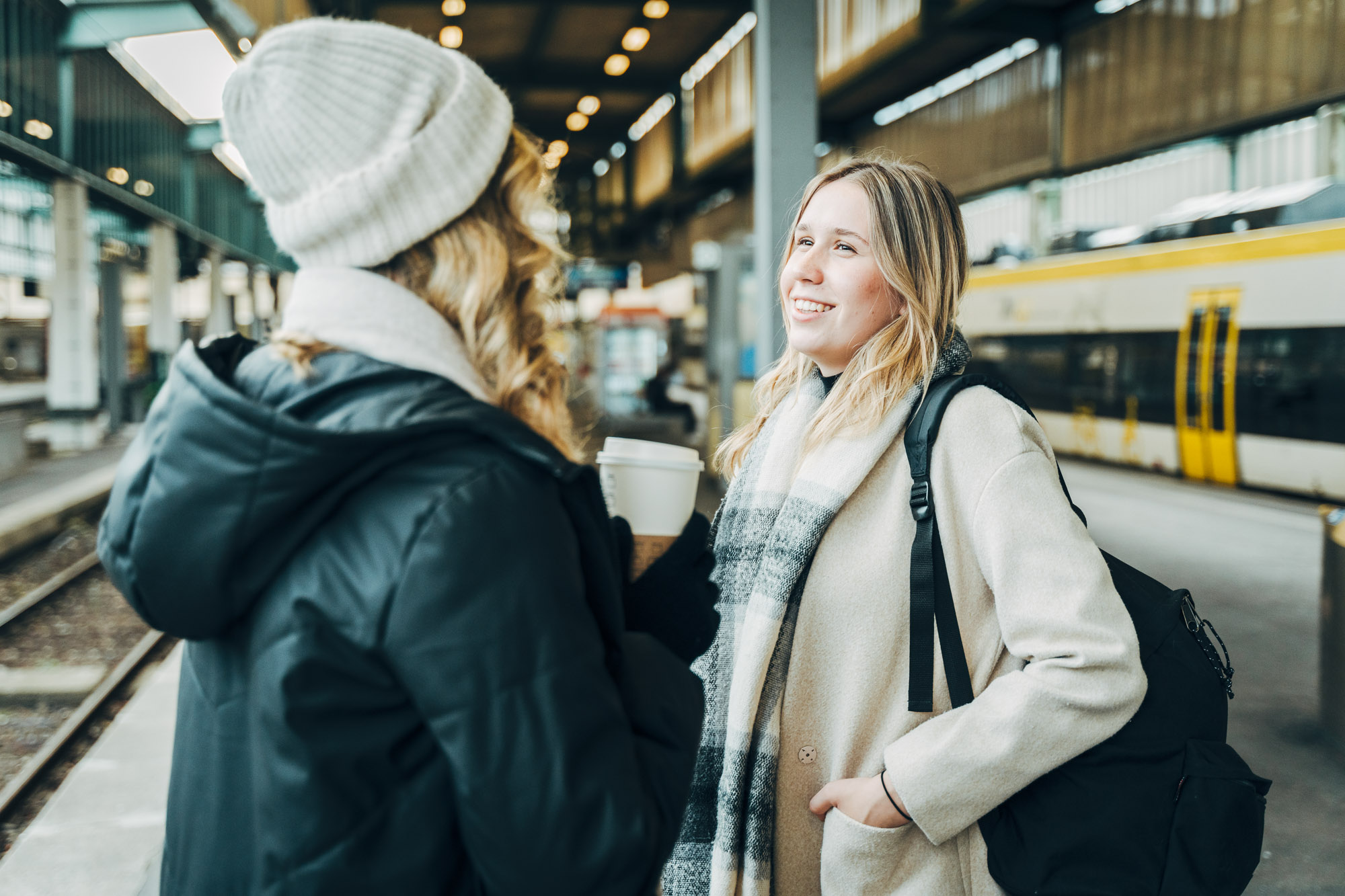 Zwei junge Frauen in warmer Winterkleidung, Schals und Mütze unterhalten sich entspannt an einem Bahnsteig. Eine der Frauen hält ein warmes Getränk in den Händen.