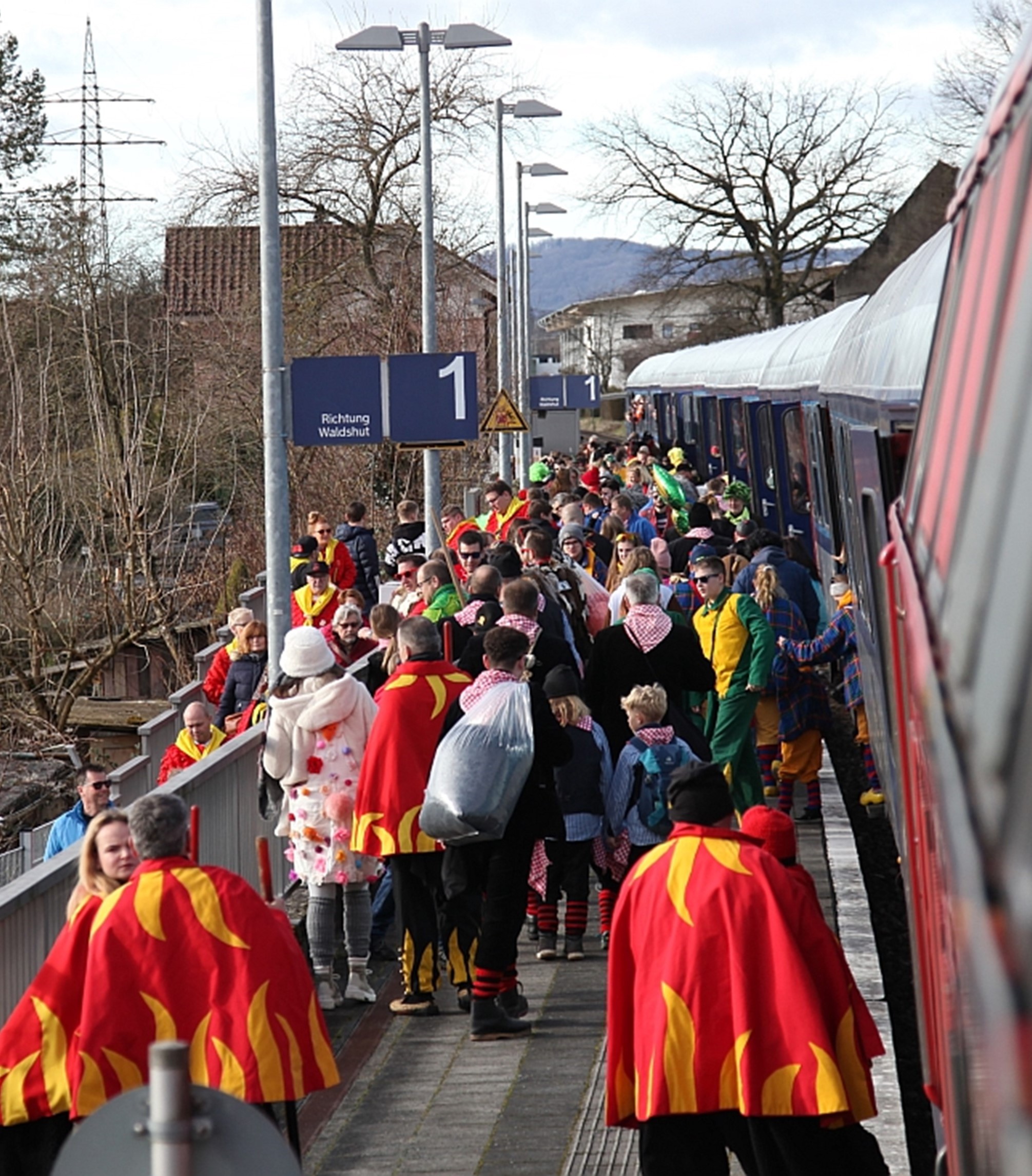 Auf einem unüberdachten Bahnsteig tummeln sich große Menschengruppen, die traditionelle süddeutsche Fastnachtskleidung tragen: rote Umhänge mit gelben Flammenspitzen. Die Menschen steigen gerade aus einem Zug. Auf einem Schild ist die Aufschrift zu lesen: „Gleis 1, Richtung Waldshut“.