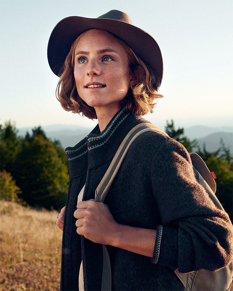 Eine junge Frau mit Wanderausrüstung und Hut ist in der Natur auf einem Feld.