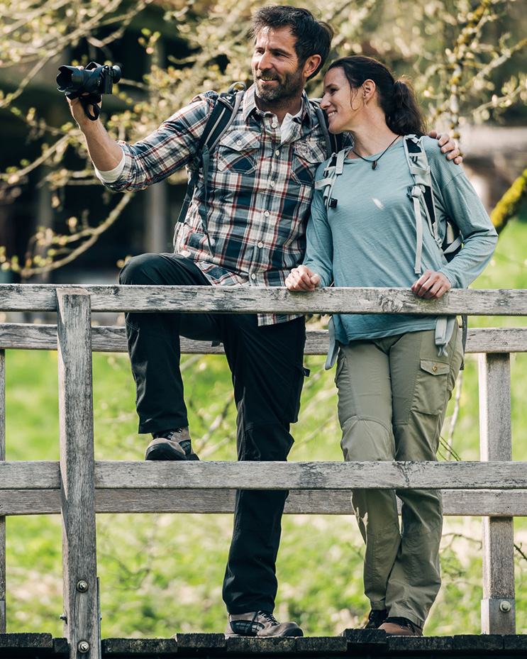 Ein Mann und eine Frau stehen auf einer Holzbrücke in der Natur. Er hat eine Kamera in der rechten Hand und fotografiert in die Ferne.